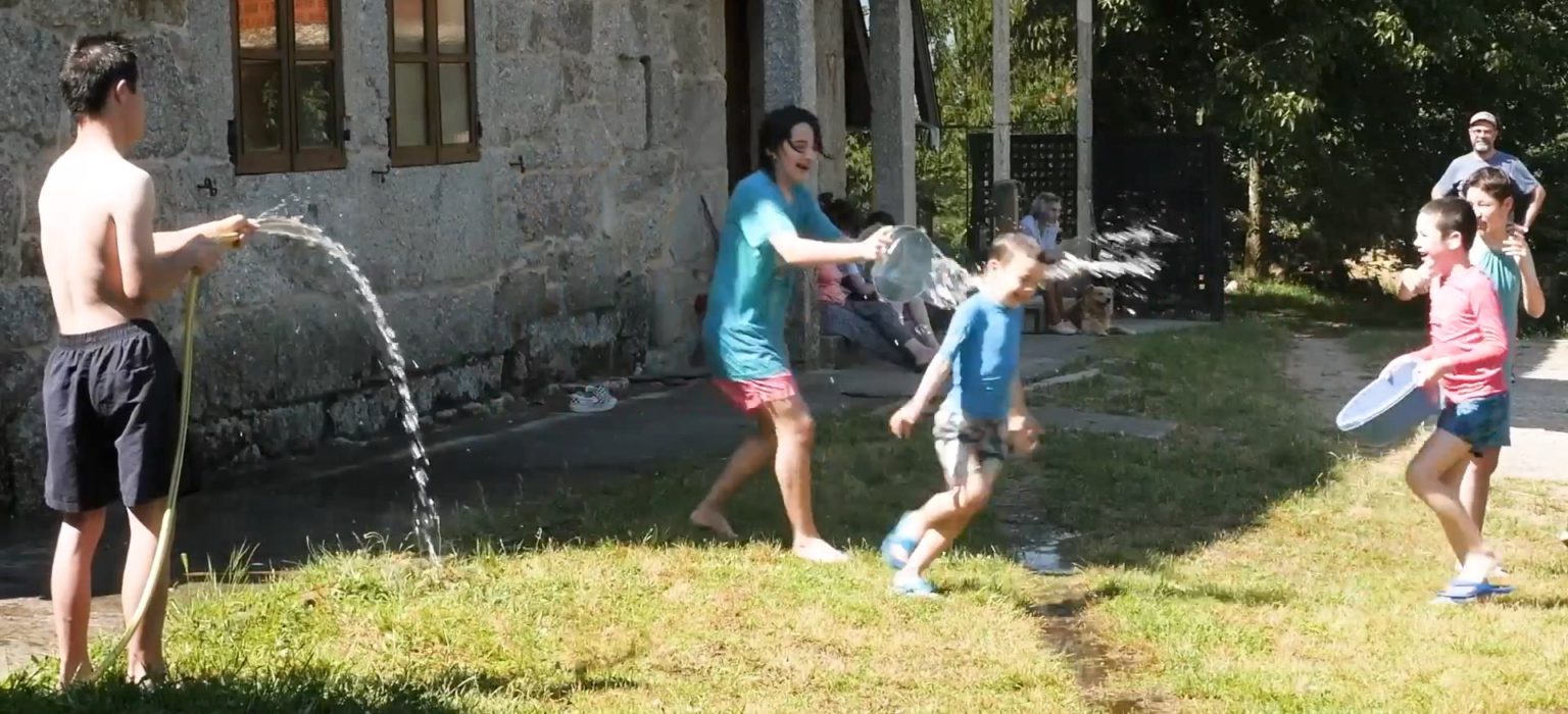 Fotografía. Vista lateral de un grupo de jóvenes jugando con agua en un jardín. Es un día soleado. A tu izquierda, una persona joven con una manguera arroja agua; en el centro, otra lanza agua desde un cubo a un niño que parece divertirse. A tu derecha, otras dos, riendo, participan con cubos. Al fondo, una persona adulta observa la escena, mientras otras conversan sentadas.