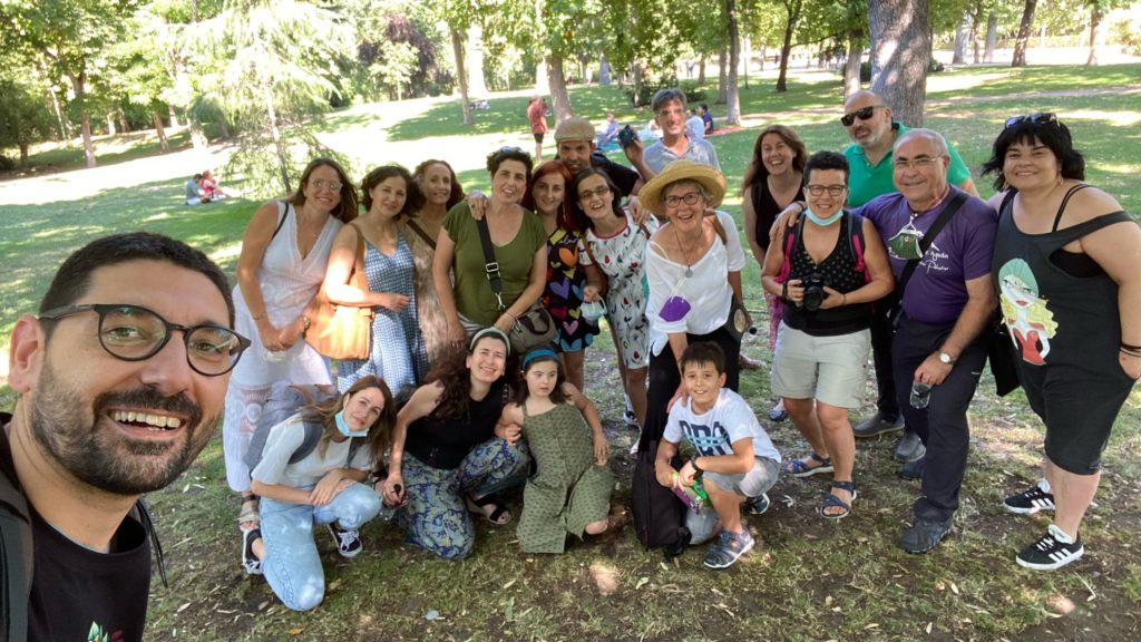 Fotografía en color. Vista frontal de un grupo de personas sonriendo en un parque. Hay personas adultas y menores, y algunas sostienen cámaras. 