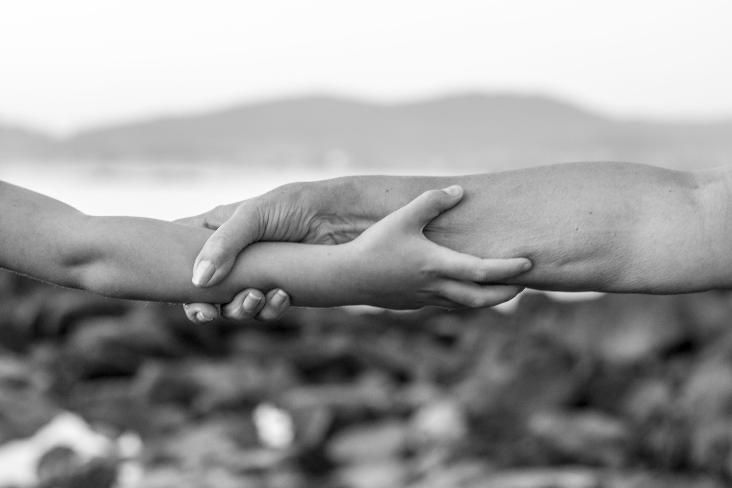 Fotografía en blanco y negro. Primer plano de dos brazos extendidos, uno de un adulto y otro de un niño, enlazados, agarrándose el antebrazo. Al fondo, un paisaje costero.