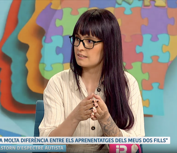 Fotograma. Entrevista en IB3. Primer plano de una mujer con gafas y cabello oscuro largo sentada en el set de televisión de IB3, decorado con un puzzle de piezas de diferentes colores. En la parte inferior, el rótulo: "Hi havia molta diferència entre els aprenentatges dels meus dos fills. 12:18. Trastorn d'espectre autista."