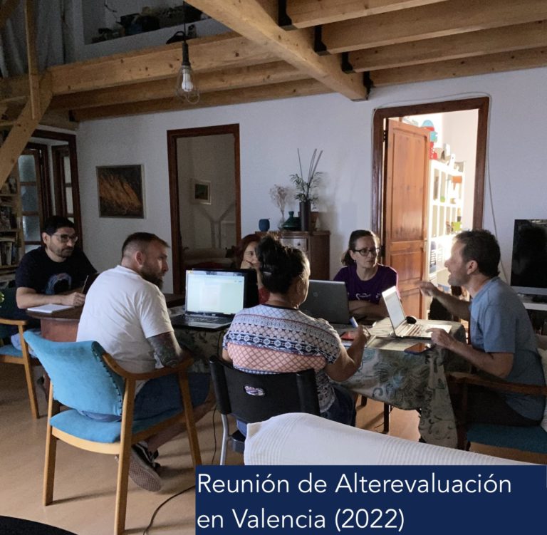 Fotografía. Reunión de Alterevaluación en Valencia (2022). Vista posterior de una reunión de grupo llevada a cabo en un estudio con vigas de madera visibles y decoración hogareña. Seis personas están sentadas alrededor de una mesa, conversando, con varios portátiles abiertos frente a ellos.