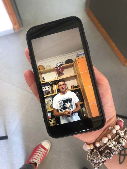 Fotografía. Vista superior de una mano sujetando un teléfono, mostrando en pantalla un joven en videoconferencia.