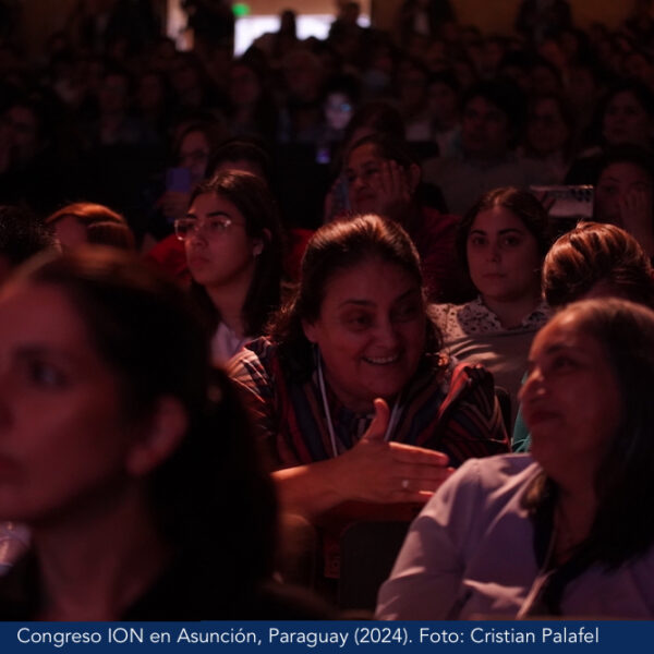 Fotografía. Congreso ION en Asunción, Paraguay en 2024. Vista general de una audiencia diversa y atenta, sentada en un auditorio. En primer plano, una mujer que sonríe e interactúa con otra persona sentada enfrente. 