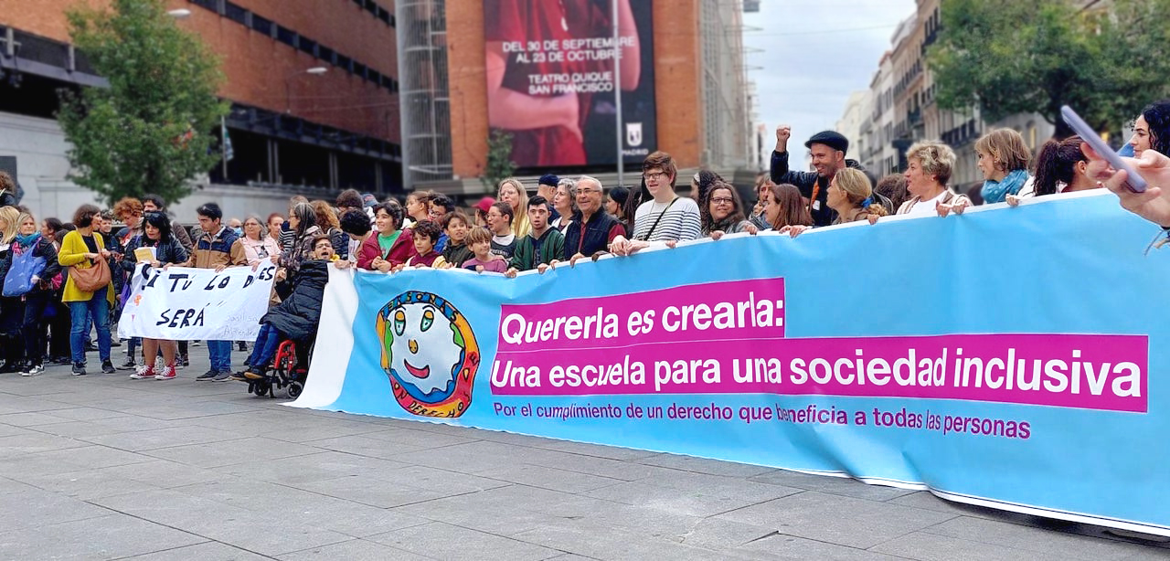 Instantánea de la manifestación «Quererla es Crearla», en la plaza Callao de Madrid. Vista lateral de un grupo de personas, jóvenes y adultas, en plena manifestación. En la cabecera de la manifestación, una lona con el lema: «Quererla es Crearla: una escuela para una sociedad inclusiva. Por el cumplimiento de un derecho que beneficia a todas las personas». Junto a ella, una pancarta más pequeña con el mensaje: «Si tú lo dices, será».