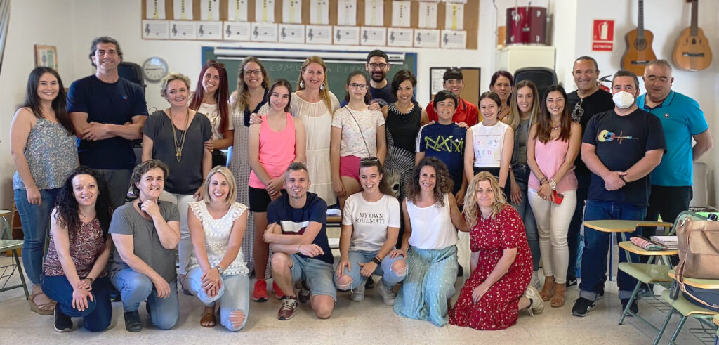 Fotografía. Aula de música. Vista frontal de 26 personas de diversas edades, participantes del grupo de acción en el centro educativo La Parra. Tras el grupo, una pizarra de tiza, una docena de pentagramas impresos y varios instrumentos musicales, como guitarras y tambores.