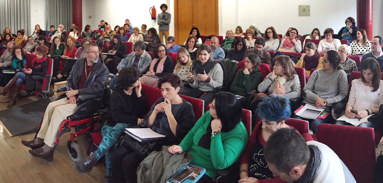 Instantánea del workshop «Orienta», realizado en Málaga. Vista lateral de un grupo de personas, con y sin discapacidad, sentadas en una sala o auditorio. Una mujer sostiene un micrófono y parece dirigirse al resto de las personas durante su intervención. A su alrededor, todo el grupo presta atención.