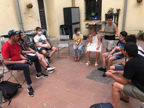 Fotografía. Detalle de un grupo de personas jóvenes y adultas sentadas en círculo en una terraza o patio.