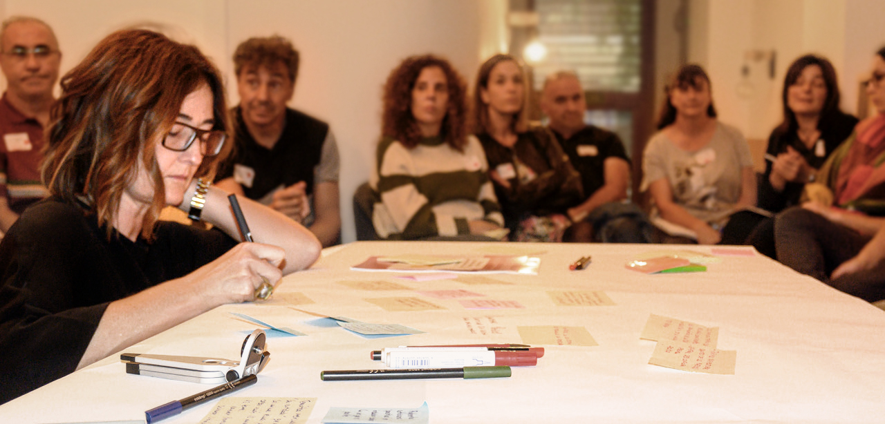 Fotografía. Instantánea del workshop «Crearla». En primer plano, una persona adulta concentrada en escribir sobre una mesa con bolígrafos de colores y notas adhesivas esparcidas. Al fondo, un grupo de participantes agrupados a su alrededor.