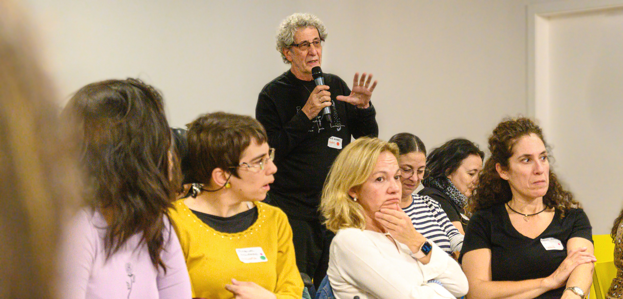 Instantánea del workshop «Crearla», realizado en Madrid. Vista lateral de un adulto, de pie, hablando con un micrófono en la mano. Parece dirigirse a alguien fuera de plano durante una intervención. A su alrededor, un grupo de personas sentadas presta atención.