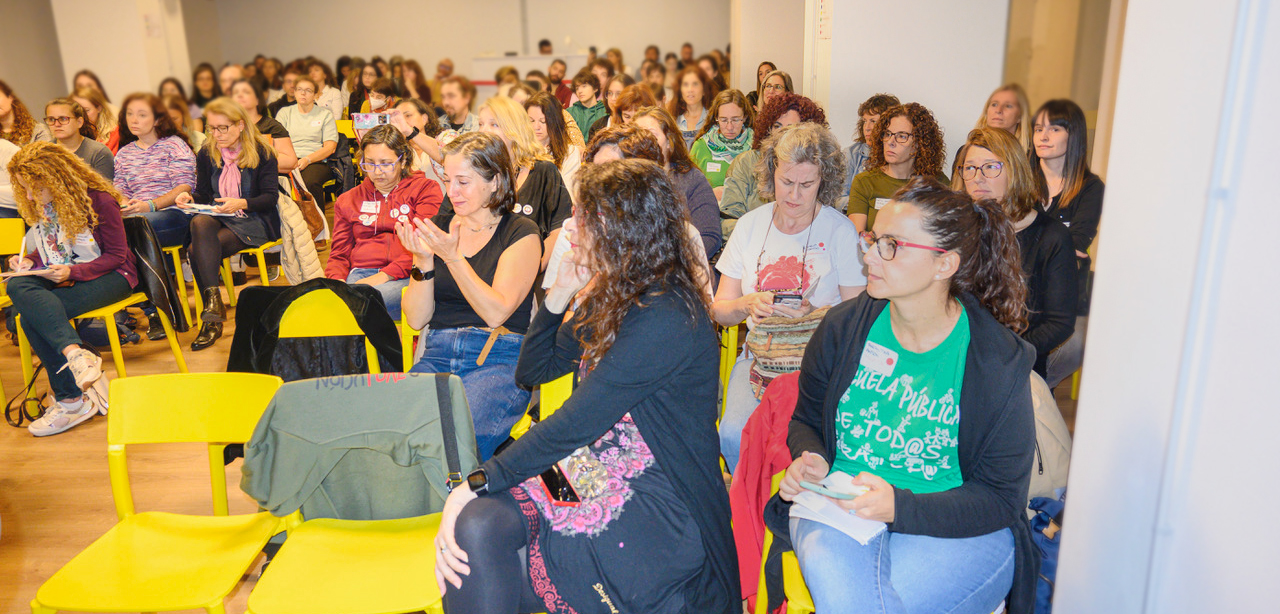 Instantánea del "Workshop Crearla", en Madrid, 2022. Vista lateral de una sala llena con un centenar de personas, todas participantes de una asamblea. Están sentadas, atentas a algo o alguien fuera del encuadre, y varias toman notas. En primer plano, una participante lleva una camiseta verde con el lema «Escuela pública de todas las personas para todas las personas».