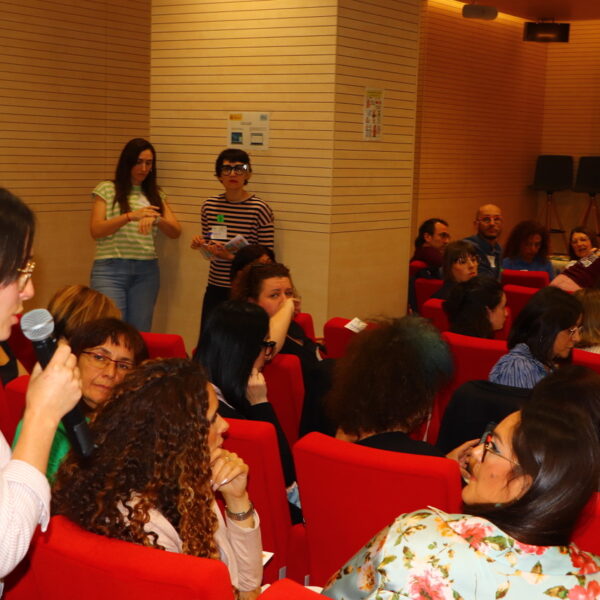 Fotografía. Mujer en pie habla con micrófono mientras otras personas escuchan sentadas, durante una asamblea en el salón de actos.