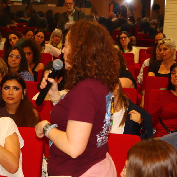 Fotografía. Mujer en pie habla con micrófono mientras otras personas escuchan sentadas.