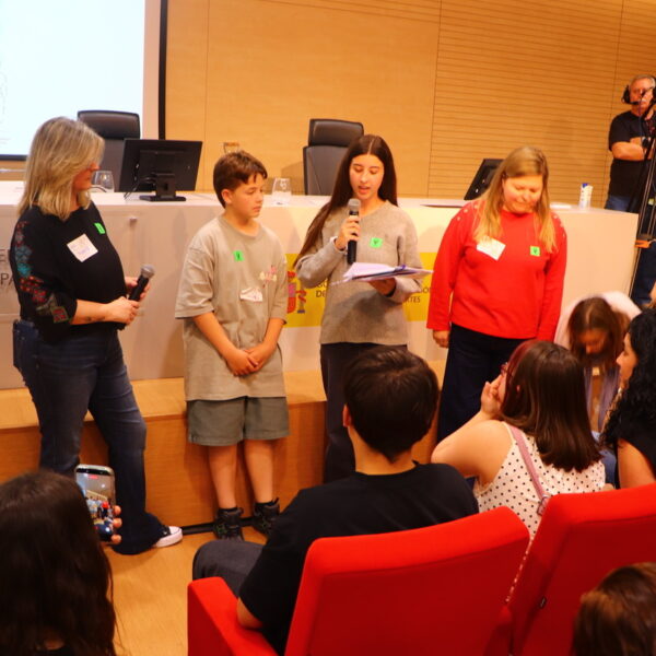Fotografía. Una joven, junto a un grupo de estudiantes, al frente, delante de la mesa del salón de actos, habla con micrófono para trasladar las ideas principales de su taller a la asamblea.