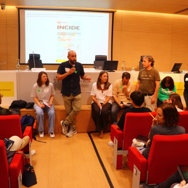 Fotografía. Un hombre, junto a otros participantes, al frente, delante de la mesa del salón de actos, habla con micrófono para trasladar las ideas principales de su taller a la asamblea.