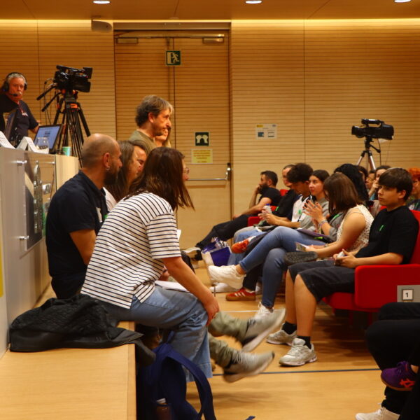 Fotografía. Varias personas sentadas al frente, delante de la mesa, durante una asamblea en el Salón de actos. Un participante toma la palabra en pie.