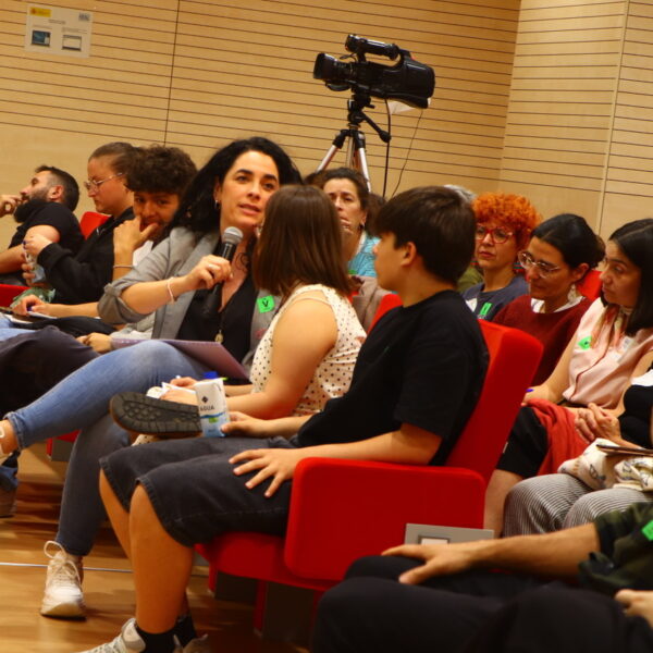 Fotografía. Mujer sentada habla con micrófono mientras otras personas escuchan sentadas, durante una asamblea en el Salón de actos.