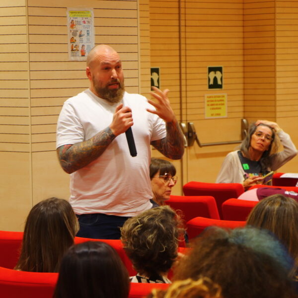 Fotografía. Hombre en pie habla con micrófono mientras otras personas escuchan sentadas durante una asamblea en el Salón de actos.