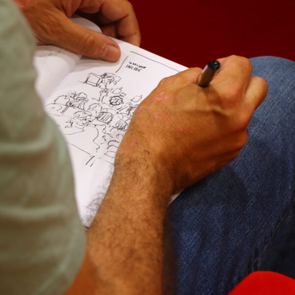Fotografía. Sobre las piernas de un hombre, un cuaderno abierto en el que está haciendo un dibujo de la situación durante una asamblea en el salón de actos.