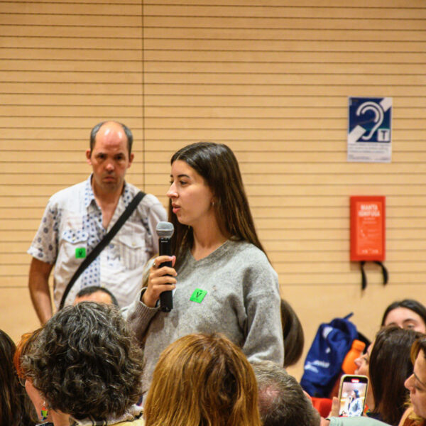 Fotografía. Mujer joven en pie habla con micrófono mientras otras personas escuchan sentadas.
