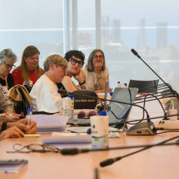 Fotografía. Un grupo de personas, sentada en una mesa de reuniones con micrófonos, escucha y toma notas durante un taller.