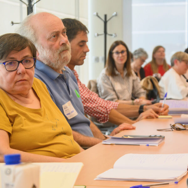 Fotografía. Un grupo de personas, sentada en una mesa de reuniones con micrófonos, escucha y toma notas durante un taller.