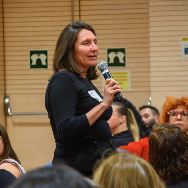 Fotografía. Mujer en pie habla con micrófono mientras otras personas escuchan sentadas, durante una asamblea en el salón de actos.