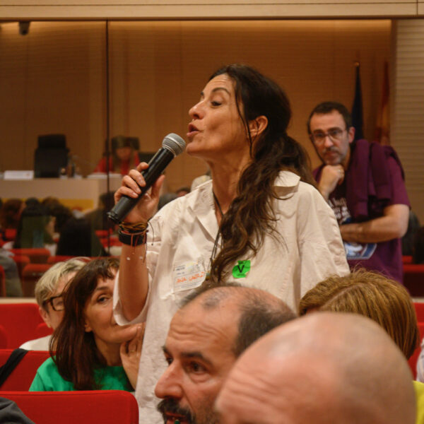 Fotografía. Mujer en pie habla con micrófono mientras otras personas escuchan sentadas, durante una asamblea en el salón de actos..