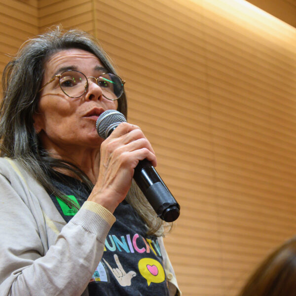 Fotografía. Mujer en pie habla con micrófono mientras otras personas escuchan sentadas, durante una asamblea en el salón de actos.
