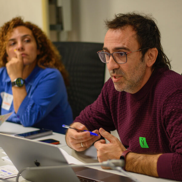 Fotografía. Participante, frente a un ordenador, toma la palabra ante la mirada de otra participante.