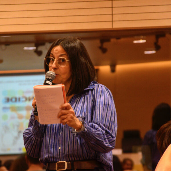 Fotografía. Mujer en pie habla con micrófono mientras otras personas escuchan sentadas, durante una asamblea en el salón de actos.