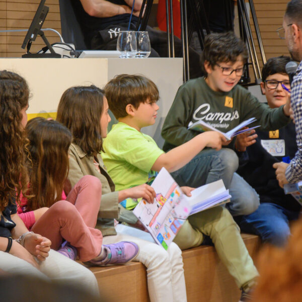 Fotografía. Un niño, junto a un grupo de estudiantes, al frente, delante de la mesa del salón de actos, habla con micrófono durante una asamblea.