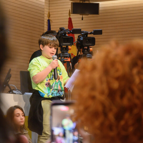 Fotografía. Un niño, junto a un grupo de estudiantes, al frente, delante de la mesa del salón de actos, habla con micrófono durante una asamblea.