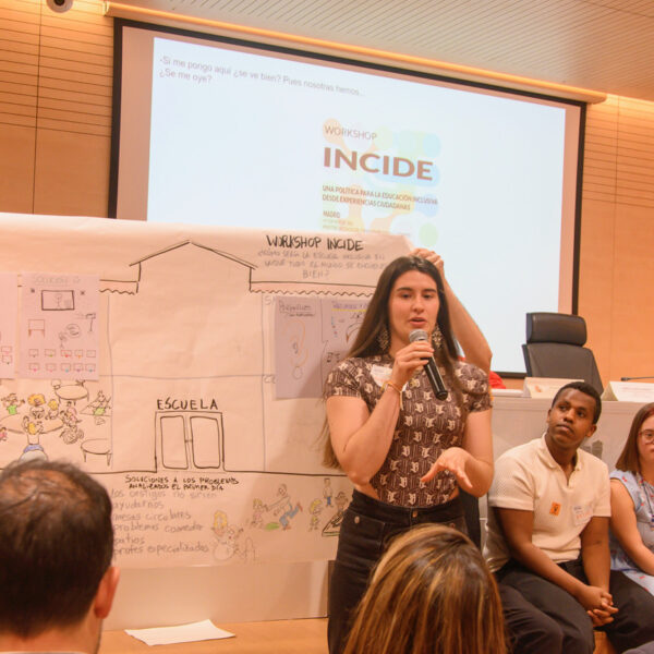 Fotografía. Una joven, junto a un grupo de estudiantes jóvenes, al frente, delante de la mesa del salón de actos y con un papelógrafo detrás, habla con micrófono durante una asamblea.