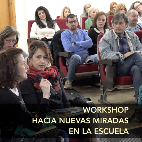 Portada de «Workshop hacia nuevas miradas en la escuela». Al fondo, una persona hablando con un micrófono. Parece dirigirse a alguien fuera de plano. A su alrededor, un grupo de personas sentadas presta atención.